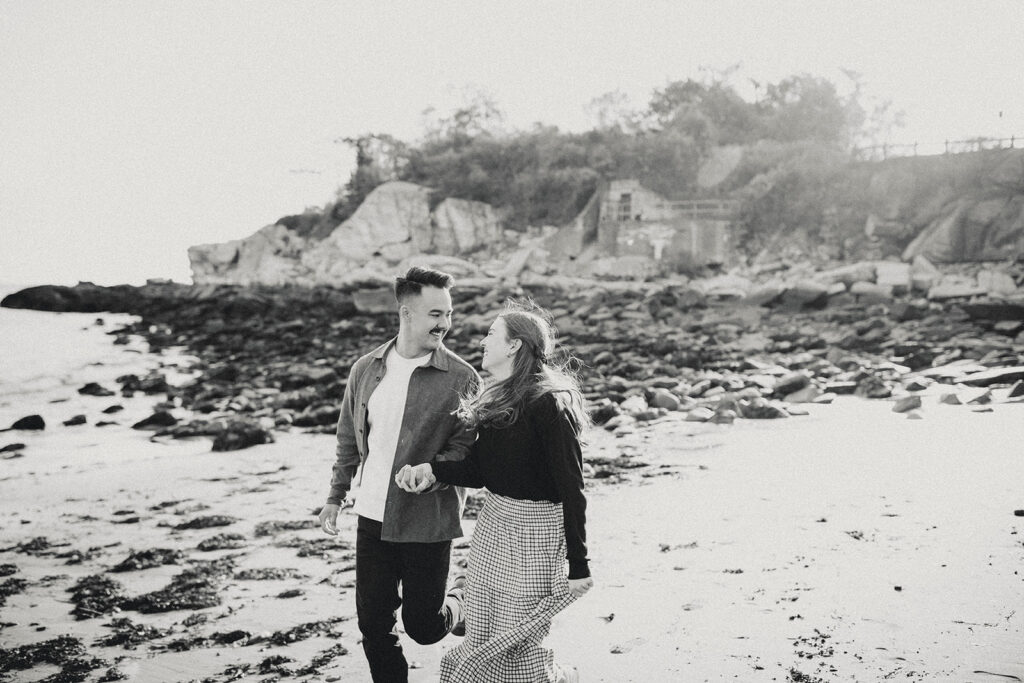 Couple holding hands and walking along rocky beach shoreline in black and white engagement photo.