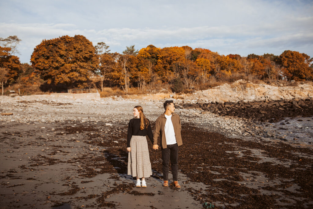 Couple holding hands while standing on seaweed-covered beach with rocky shoreline and vibrant fall trees behind them.