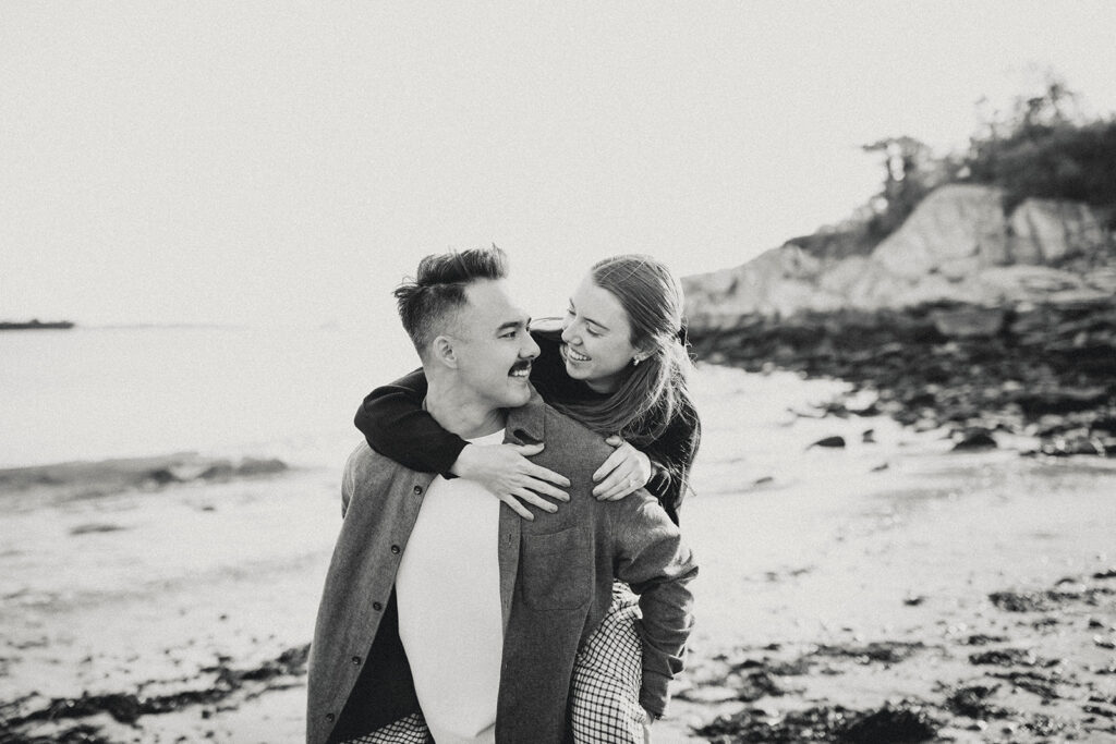 Woman hugs partner from behind while he smiles as they stand together on rocky shoreline in black and white engagement portrait.
