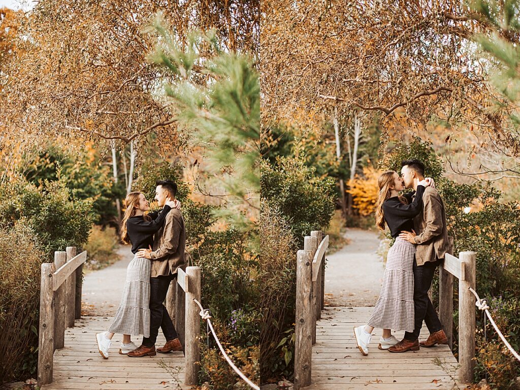 Couple kissing while standing on a small wooden bridge surrounded by fall foliage and greenery in a garden setting.