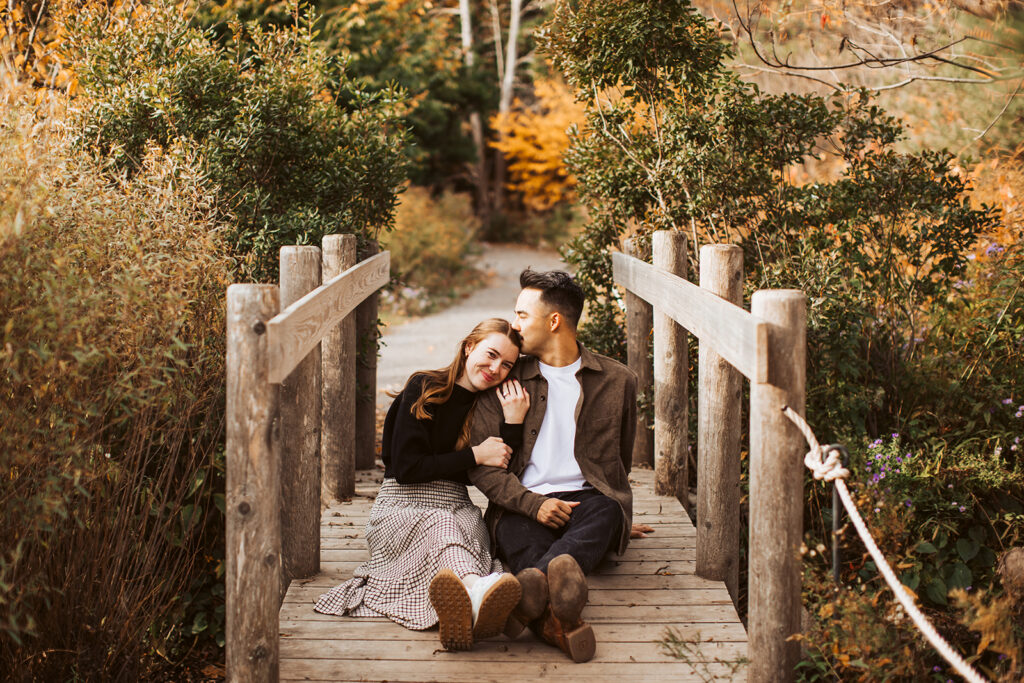 Couple sitting together on wooden bridge while woman rests her head on partner’s shoulder in warm autumn light.