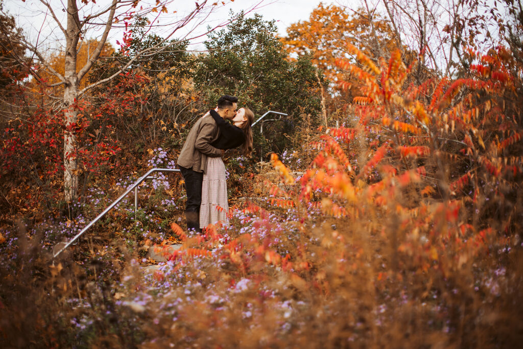 Couple kissing on a garden path surrounded by colorful fall foliage, trees, and wildflowers during engagement session.