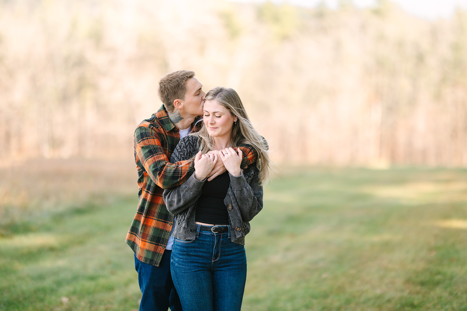 Man hugging woman from behind and kissing her head while standing in a grassy field with soft trees in background.