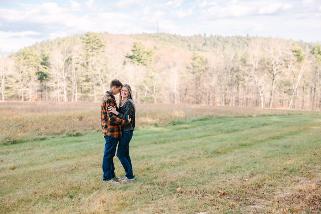 Couple standing together in wide open meadow with trees and hills in background under a partly cloudy sky.