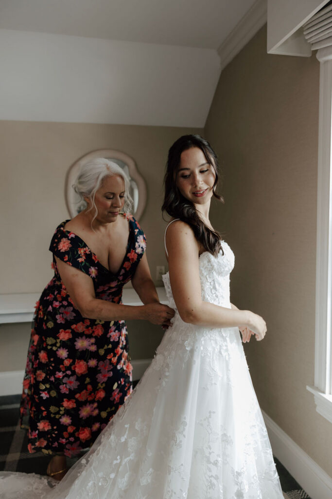Bride smiling as woman fastens back of lace wedding gown by window, soft natural light