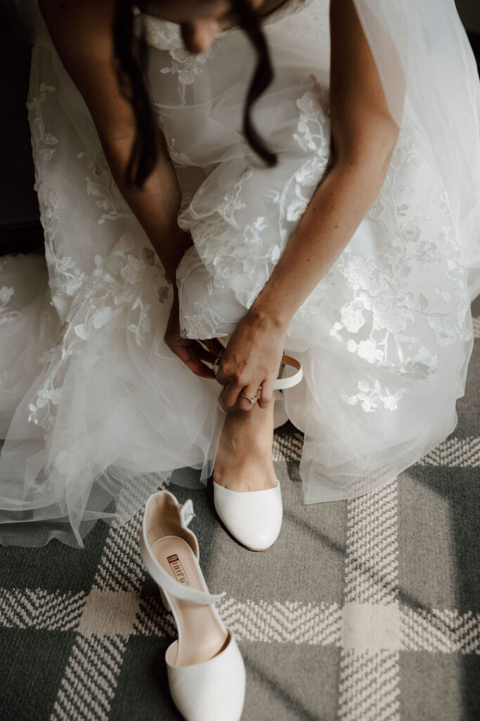 Bride fastening white wedding shoe beneath lace dress, intimate getting ready detail