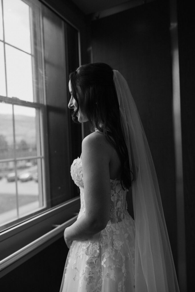 Bride in veil standing by window in black and white, quiet pre-ceremony moment