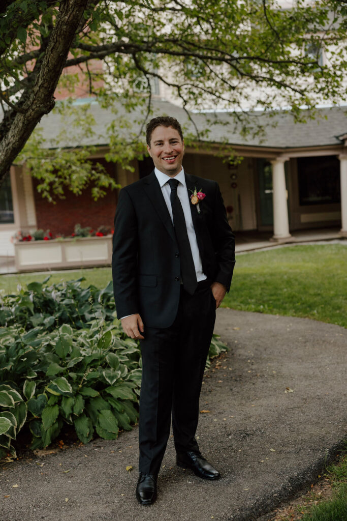 Groom in black suit with boutonniere standing outdoors near Wentworth Inn building