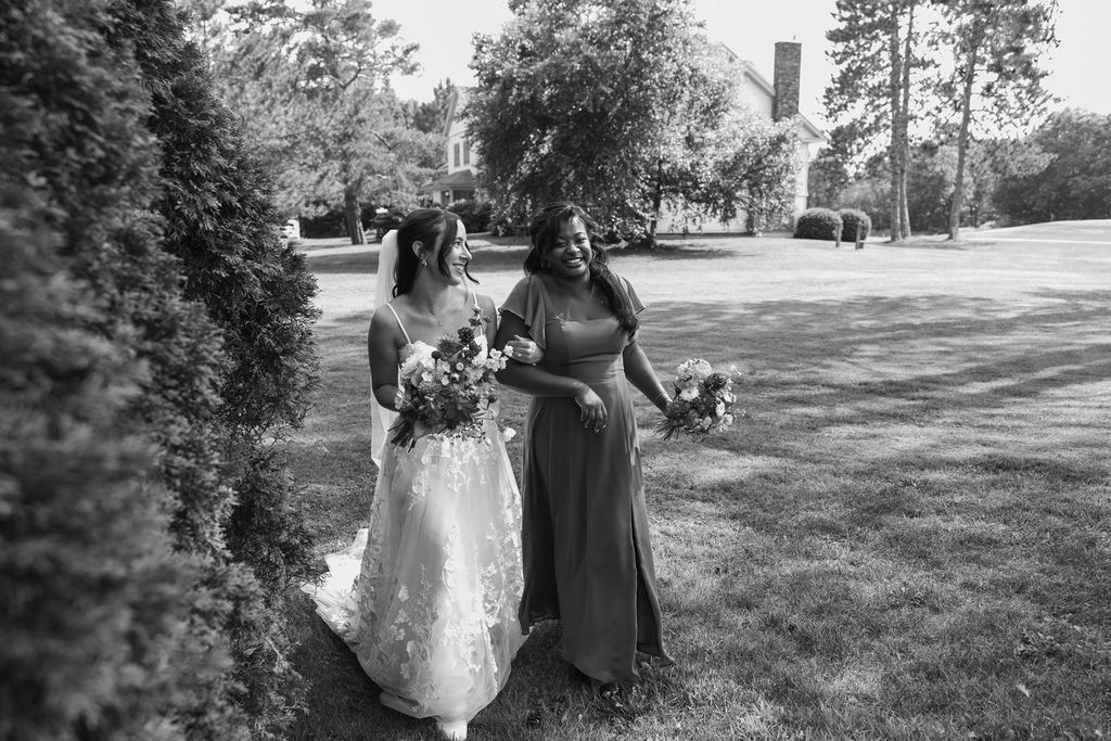 Bride and bridesmaid walking together with bouquets across lawn, candid wedding moment at Wentworth Inn