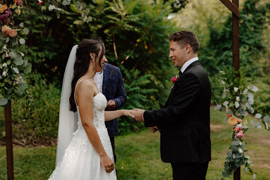 Groom placing ring on bride’s finger beneath greenery arch during wedding ceremony