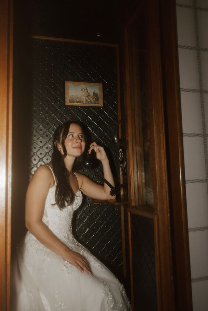 Bride in wedding dress using vintage phone booth indoors, creative bridal portrait