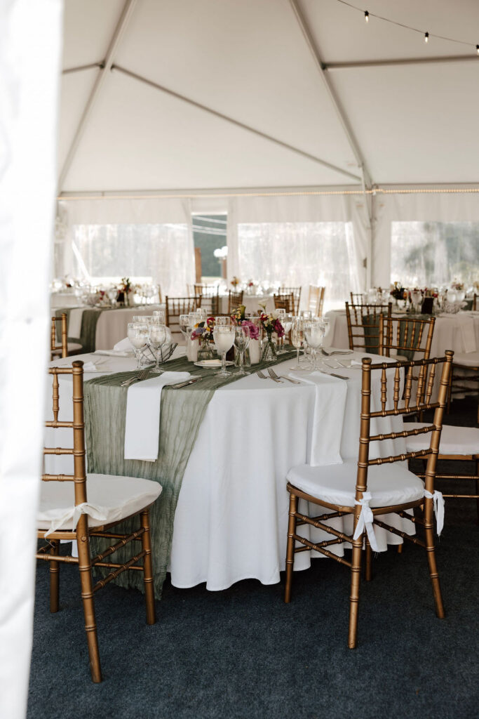 Reception tables with white linens, green runners, and gold chairs inside wedding tent
