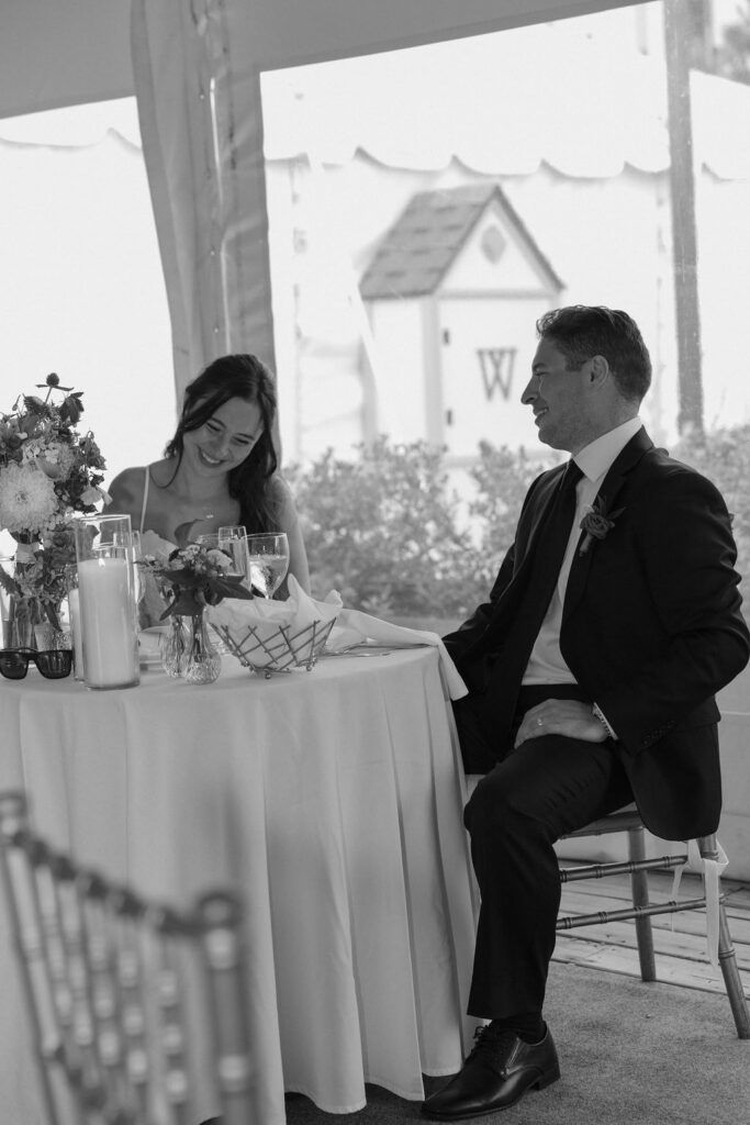 Bride and groom seated at sweetheart table laughing during reception, black and white moment