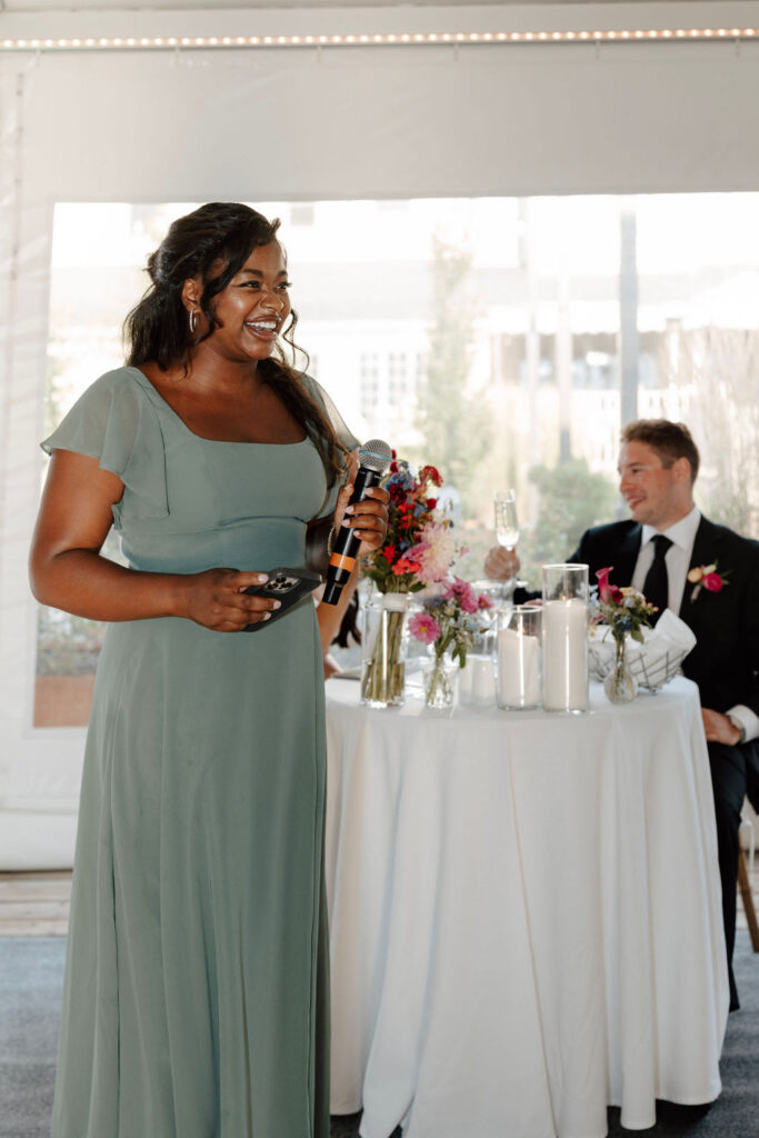 Woman giving wedding toast with microphone while groom listens at reception table