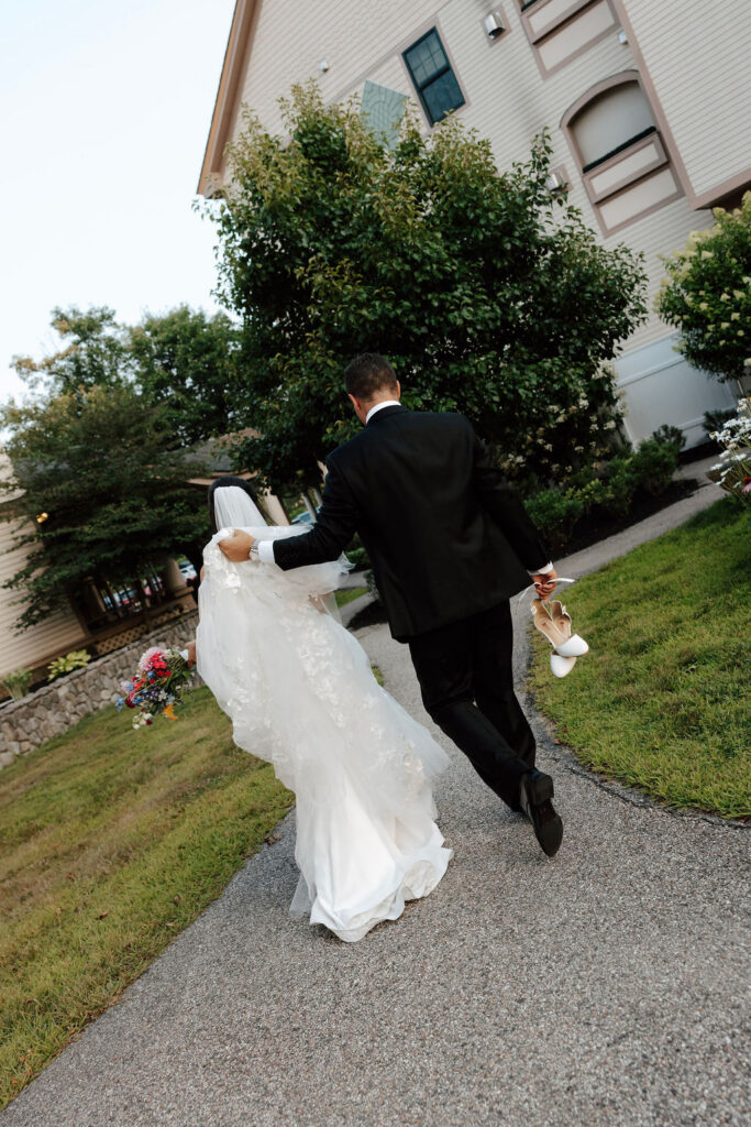 Bride and groom walk along a garden path, groom holding bride’s shoes, wedding venue in background