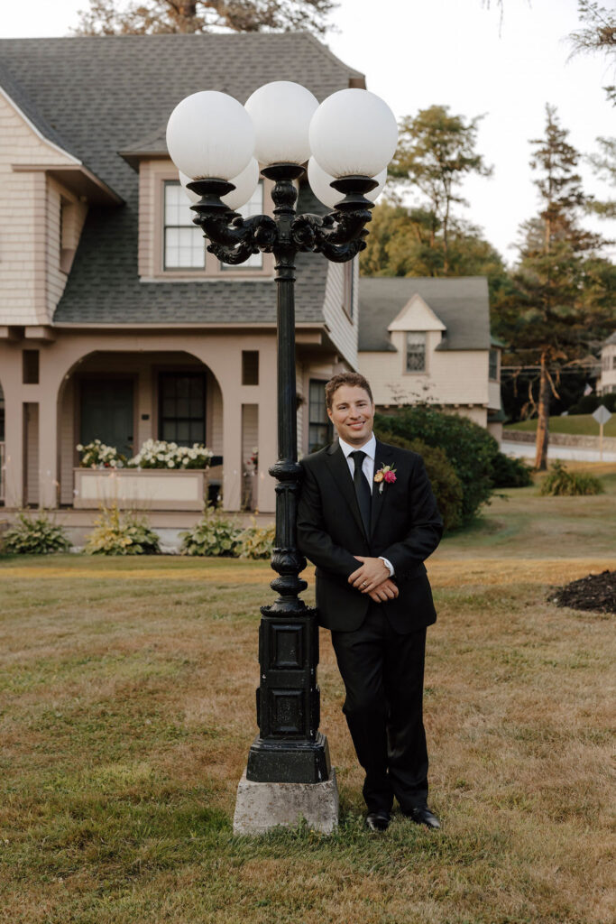 Groom in black suit leans against vintage street lamp in front of charming house at outdoor wedding venue