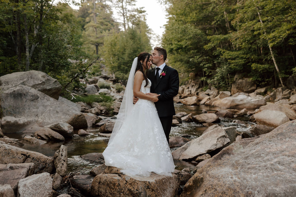 Bride and groom stand on rocks by forest stream, intimate outdoor wedding portrait in nature