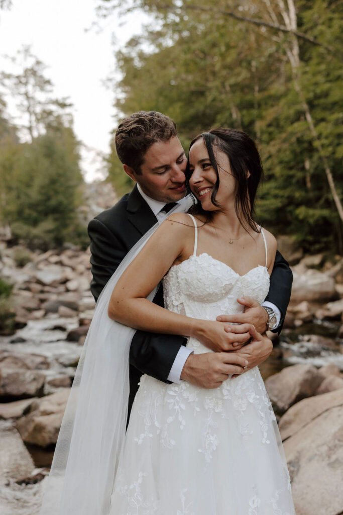 Groom embraces bride from behind by river, couple smiling during outdoor wedding portrait session