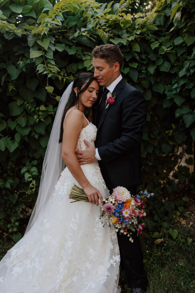 Bride and groom embrace in front of lush greenery, bride holding vibrant bouquet, garden wedding portrait
