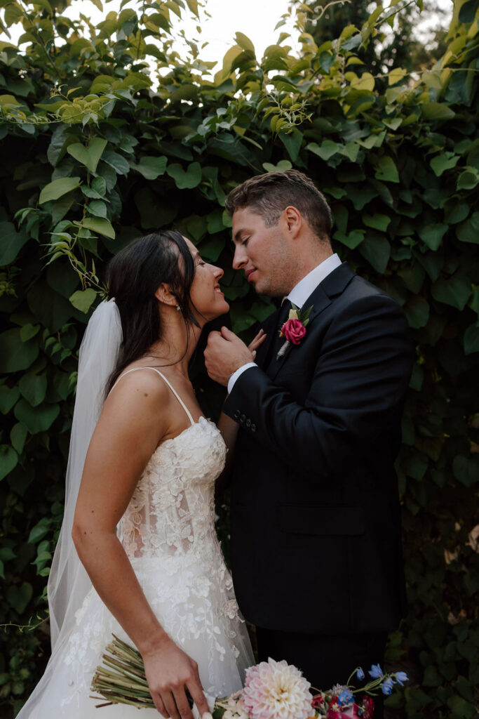 Bride and groom stand close against leafy backdrop, intimate outdoor wedding portrait with soft natural light